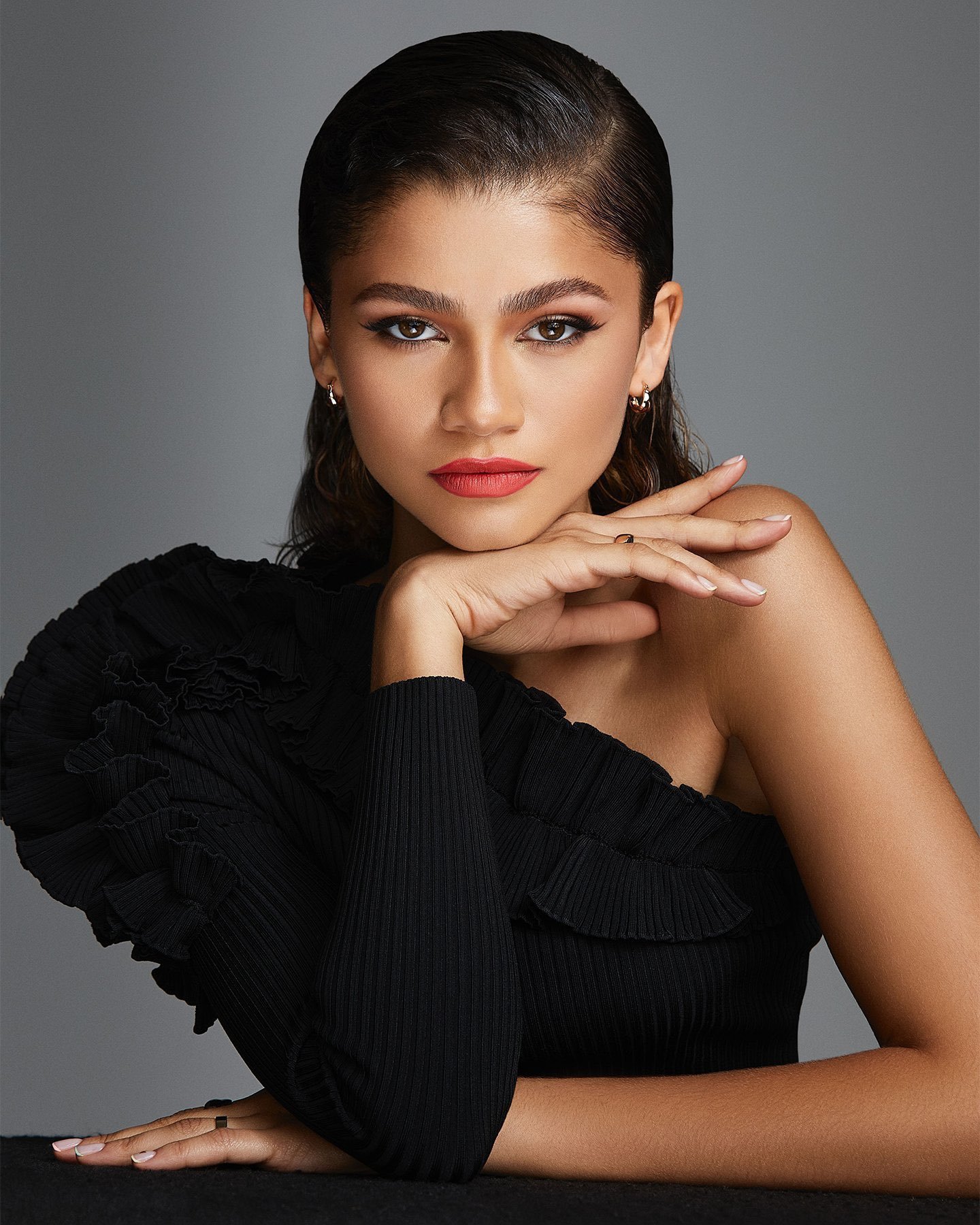Zendaya with slicked-back dark hair and red lipstick wearing a black one-shoulder top against a grey studio backdrop
