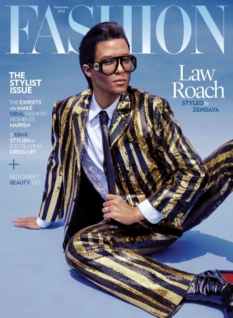Fashion magazine cover featuring Law Roach wearing a gold-striped suit and glasses, seated against a gradient blue background.