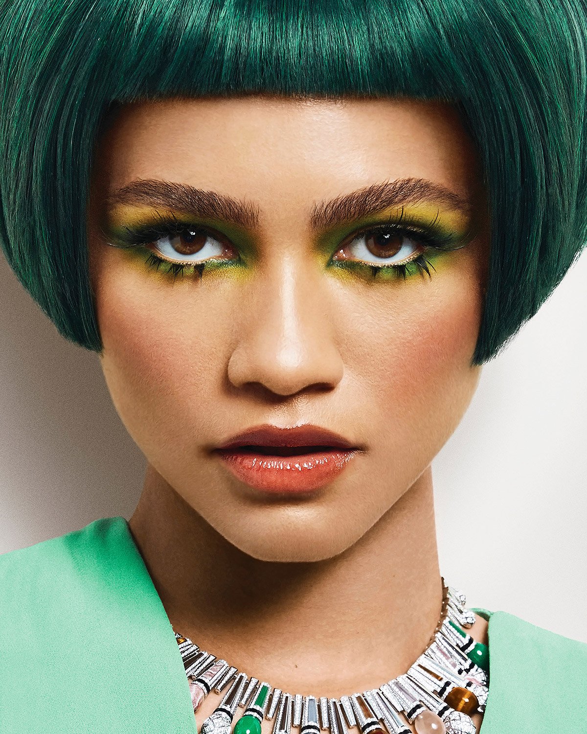 Close-up of a woman with striking green hair and vibrant yellow and green makeup, wearing a unique necklace against a white backdrop.