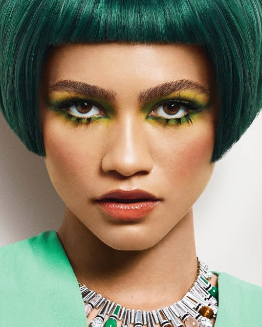 Close-up of a woman with striking green hair and vibrant yellow and green makeup, wearing a unique necklace against a white backdrop.