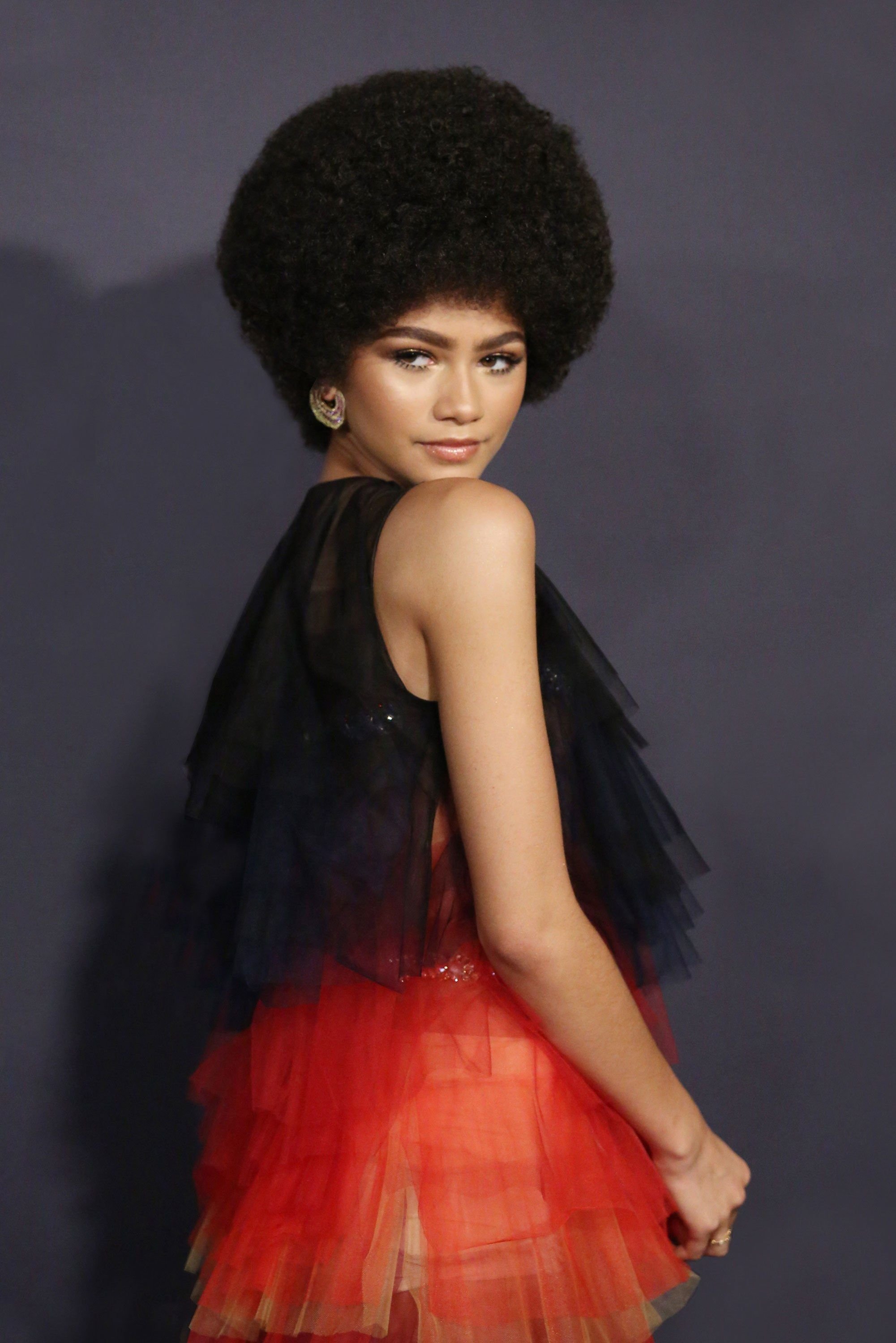 Zendaya poses in a black top with a layered red skirt and her large, dark afro hairstyle, shot in a photo studio.