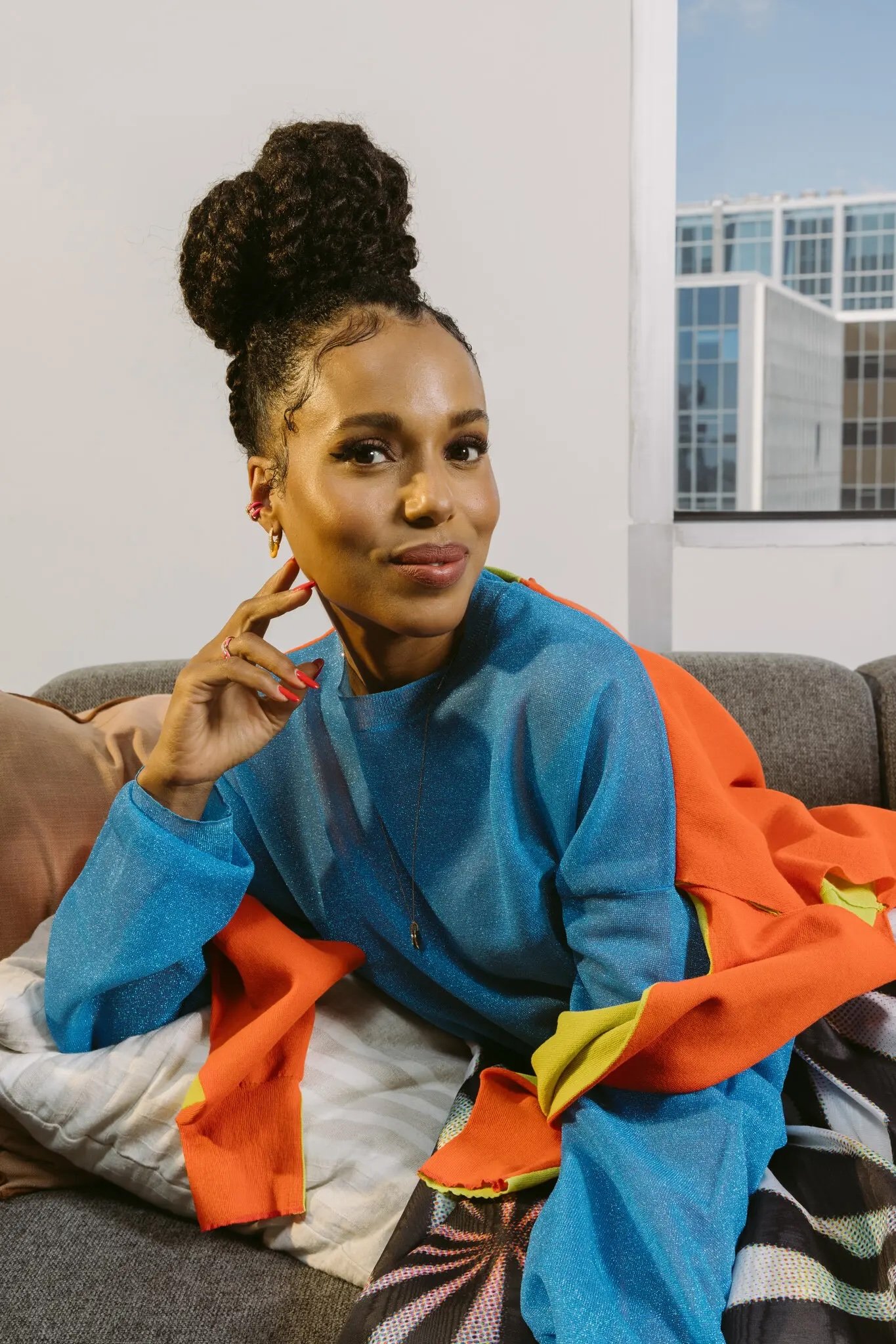Close-up shot of a smiling adult woman with a high bun posing on a grey sofa indoors with pillows and a building visible through a window.