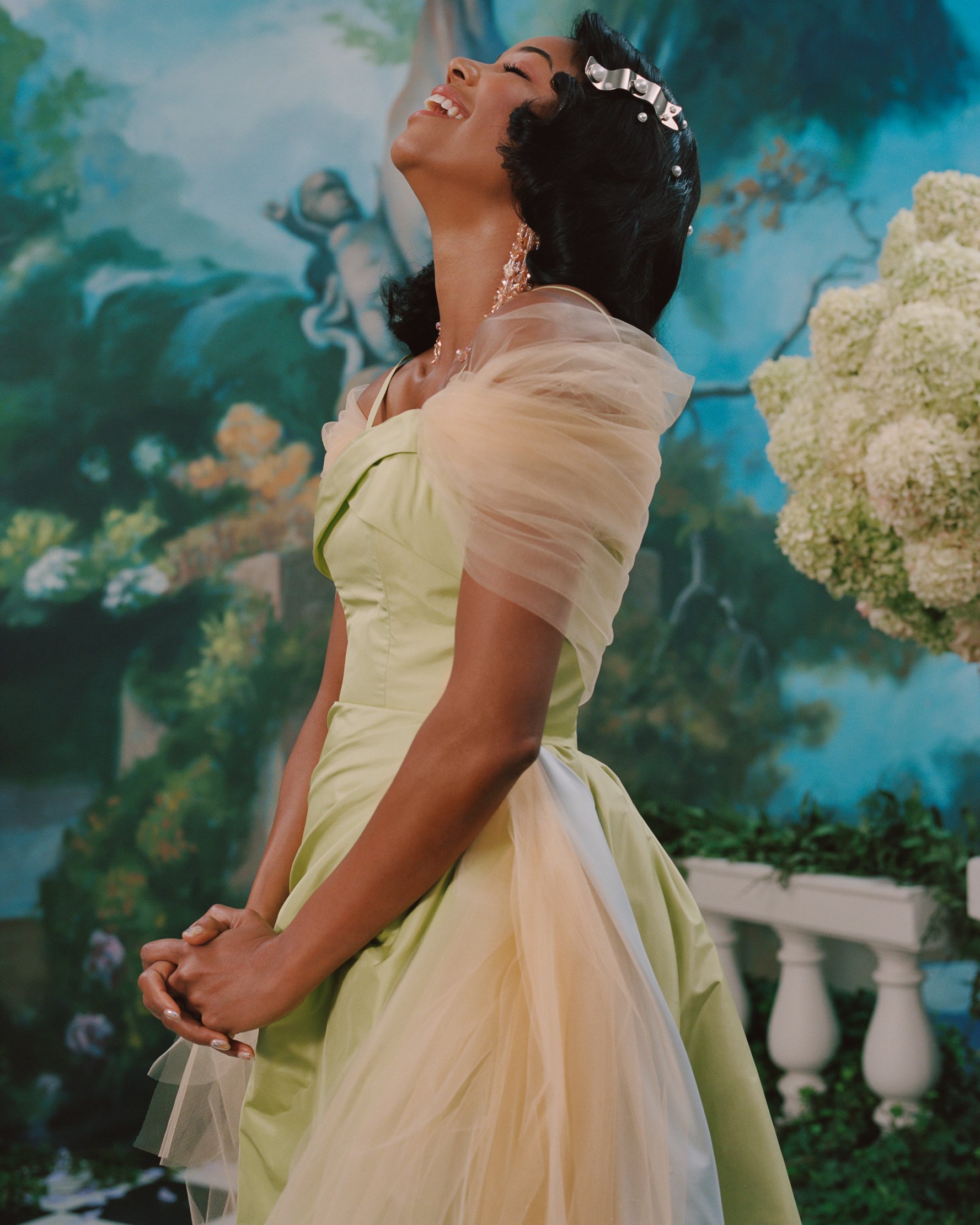 Elegant woman in a green and cream gown smiles with her head tilted back in a lighthearted moment, set against a painted backdrop.