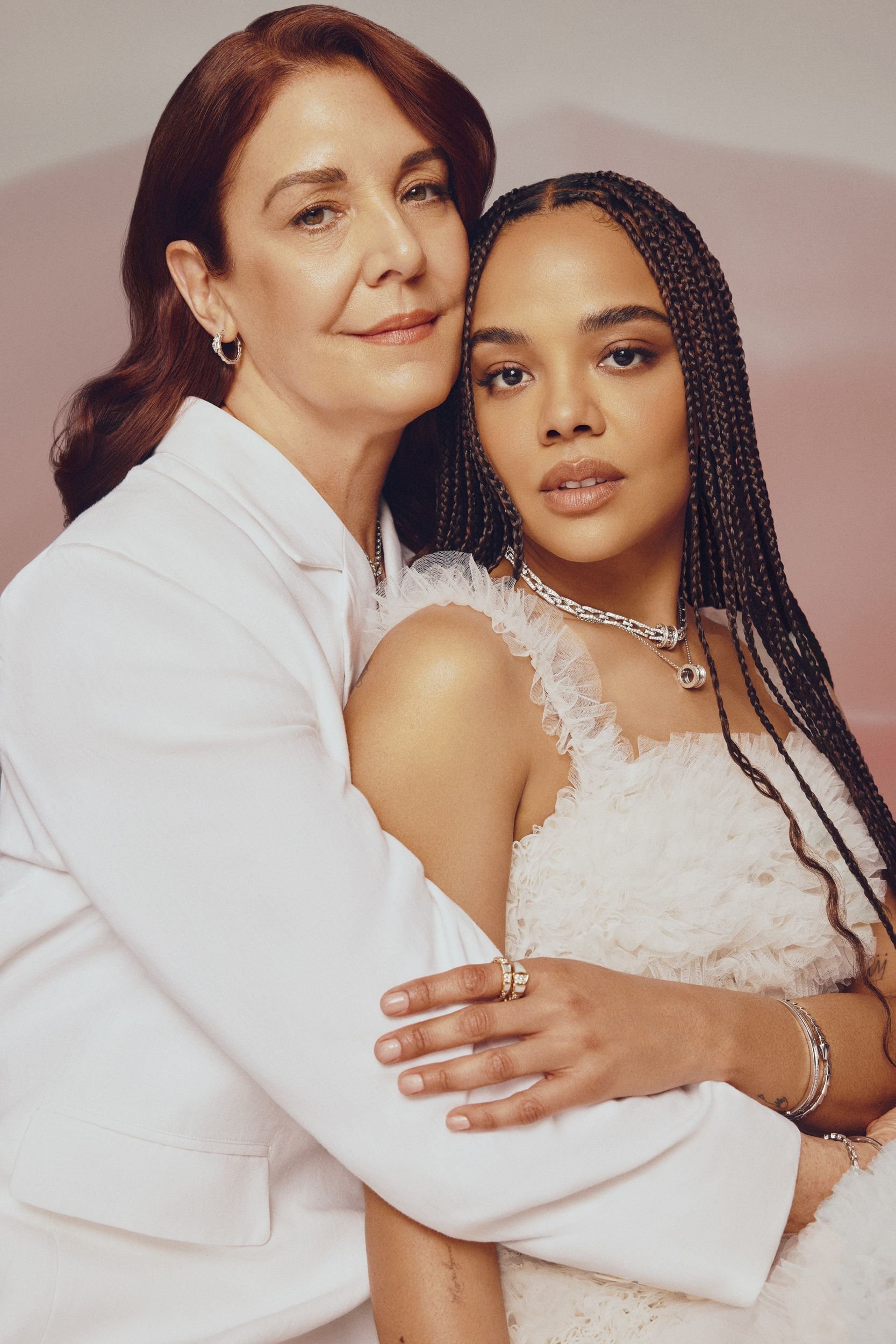 Two women in white embrace in a studio setting. One woman with auburn hair wears a blazer, while the other with dark hair has on a white gown.