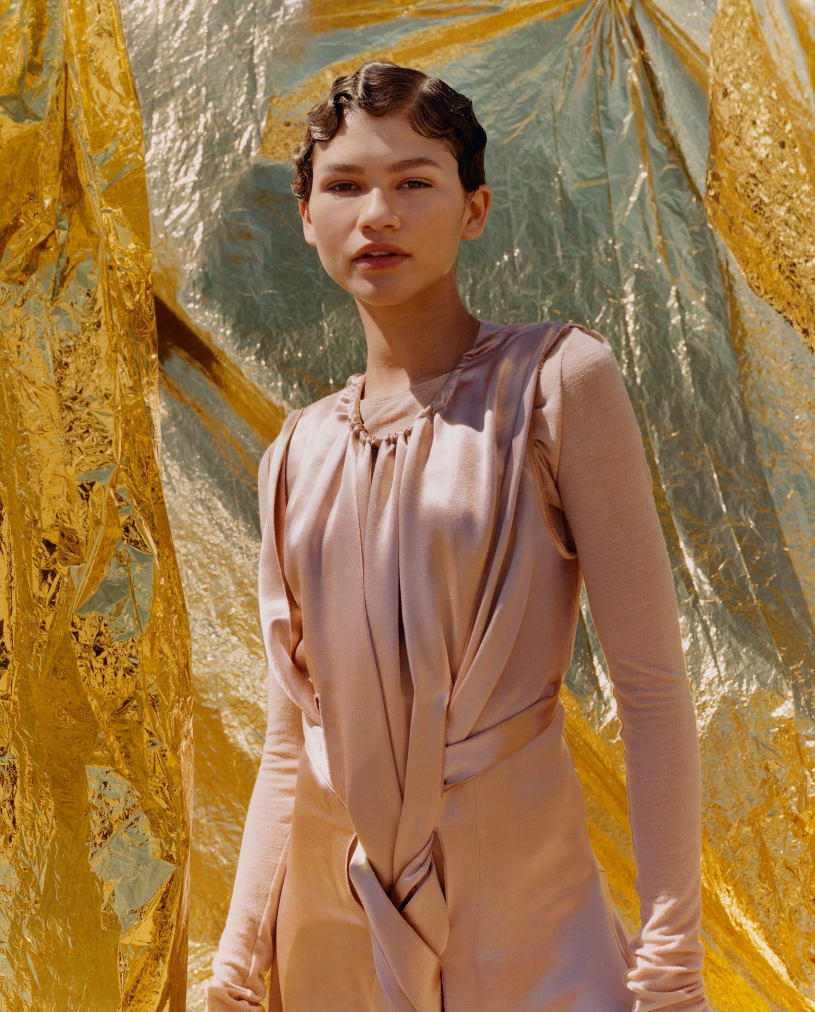 Elegant woman poses against a shimmering gold backdrop wearing a light pink gown with long sleeves and retro finger-wave hairstyle.