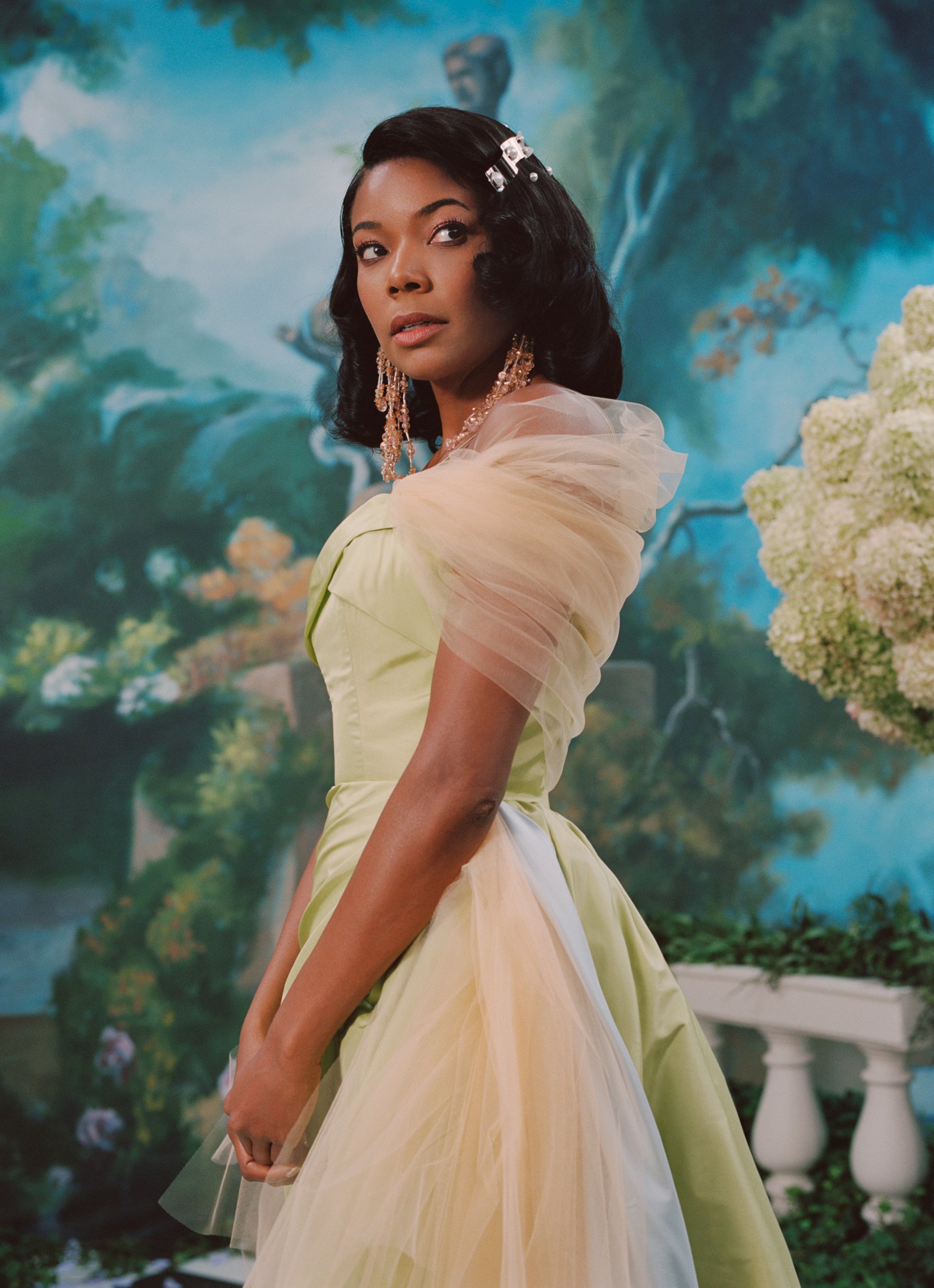 Elegant woman in a light green gown poses against a painted garden backdrop. She wears chandelier earrings and a vintage hair clip.