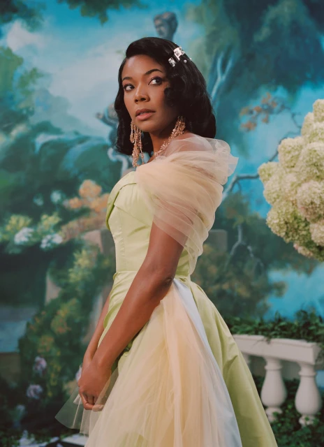 Elegant woman in a light green gown poses against a painted garden backdrop. She wears chandelier earrings and a vintage hair clip.