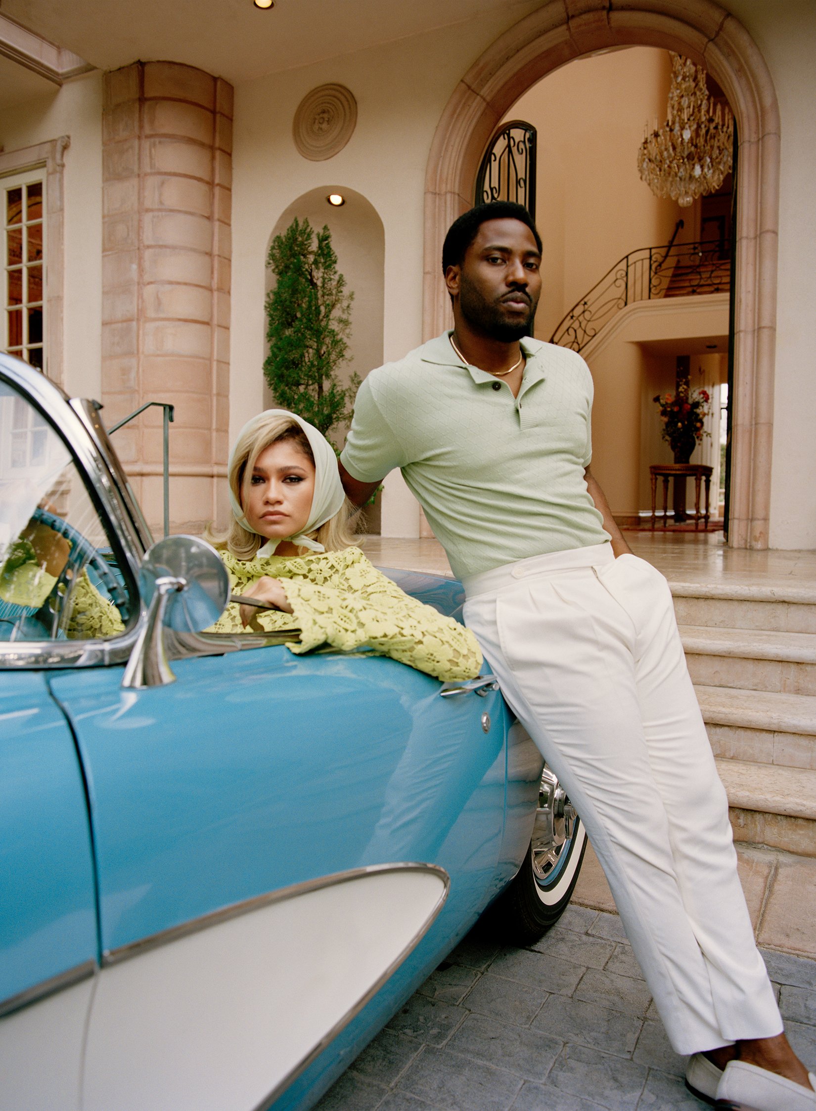 Zendaya leans from a light-blue classic car wearing a headscarf while John David Washington poses in front of a mansion, both in natural light.