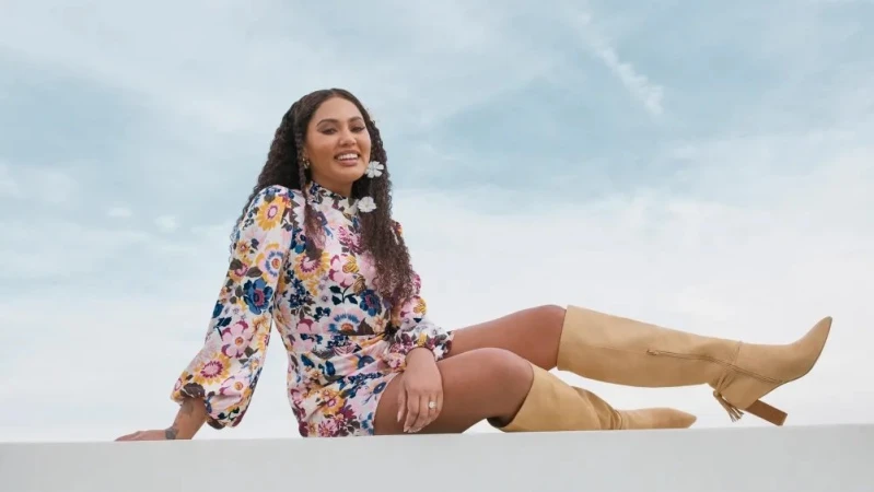 Woman with long curly hair wearing a floral dress and tan boots seated against a pale blue sky background