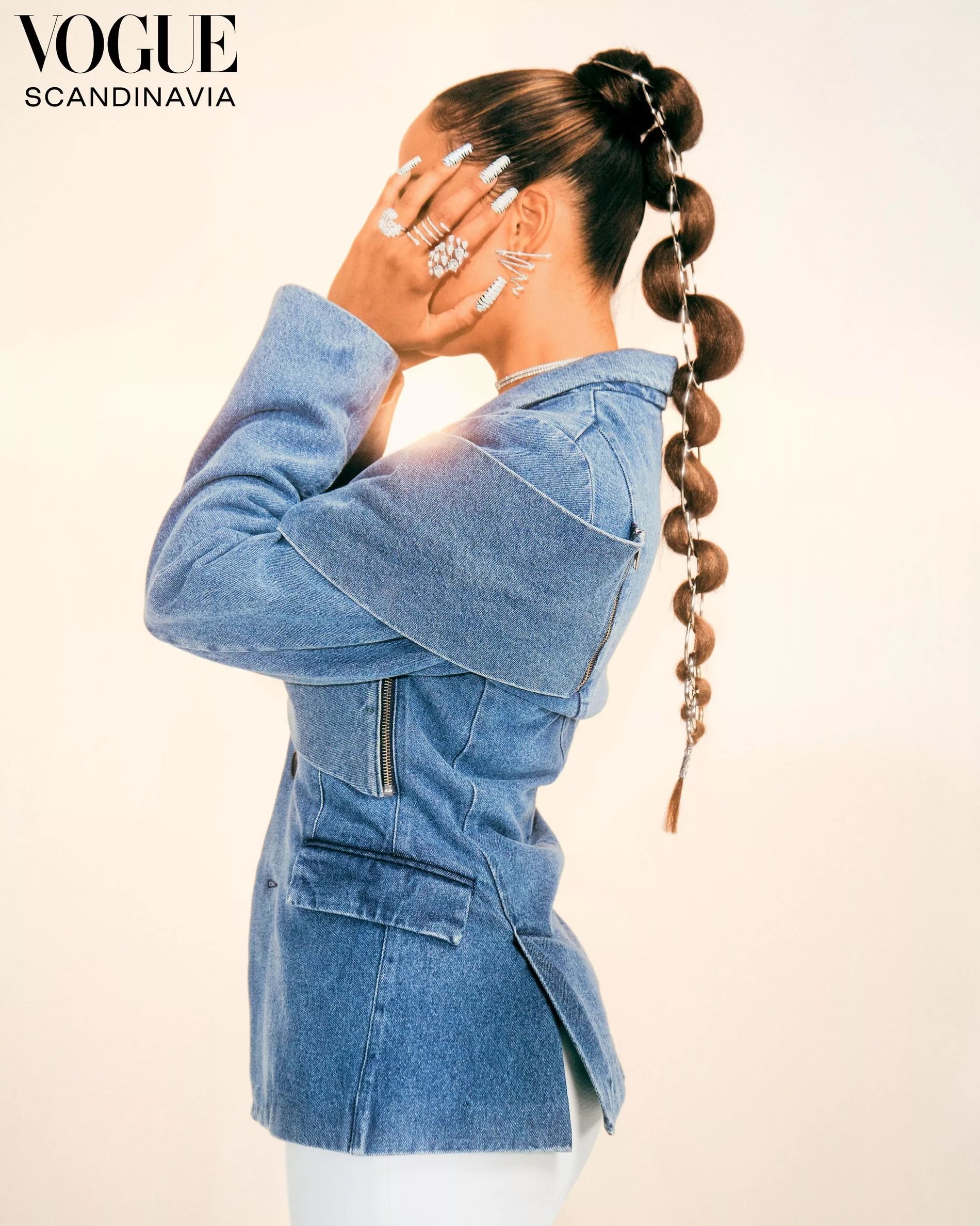 Woman in denim jacket covers her face, showcasing manicure and jewelry. Ponytail with hair-tie details and Vogue Scandinavia logo.