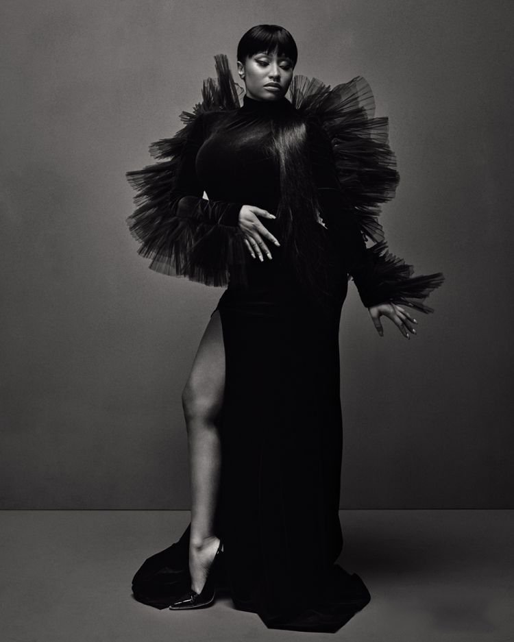 Black and white studio portrait of a woman in a gown with a high slit, accessorized with a large tulle collar and high heels.