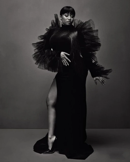 Black and white studio portrait of a woman in a gown with a high slit, accessorized with a large tulle collar and high heels.