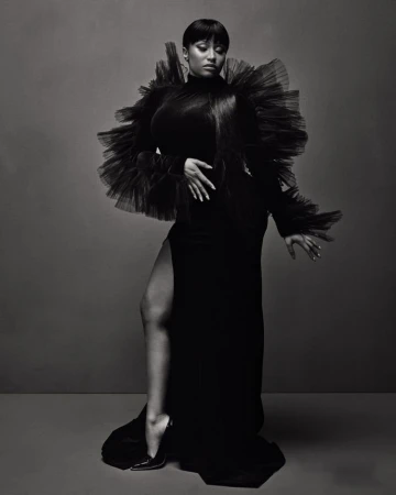 Black and white studio portrait of a woman in a gown with a high slit, accessorized with a large tulle collar and high heels.