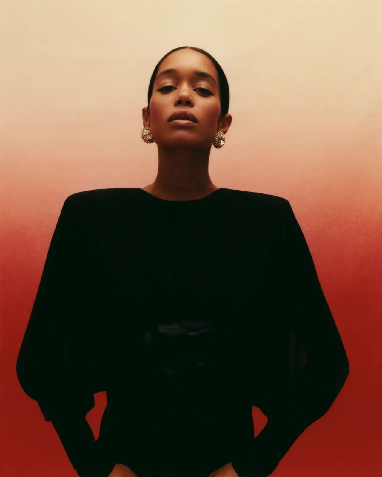 Portrait of a black female fashion model wearing a black dress with shoulder pads and pearl earrings, posing against an orange gradient background.