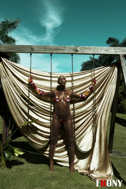 Adult female in swimwear poses on a swing against a background of blue sky and tropical trees on a sunny day outdoors.