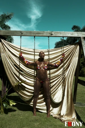 Adult female in swimwear poses on a swing against a background of blue sky and tropical trees on a sunny day outdoors.