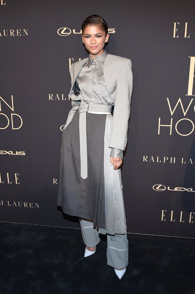 Zendaya poses in grey belted coat and wide-leg trousers at a fashion event; dark background with brand logos.