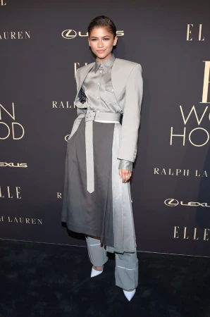 Zendaya poses in grey belted coat and wide-leg trousers at a fashion event; dark background with brand logos.