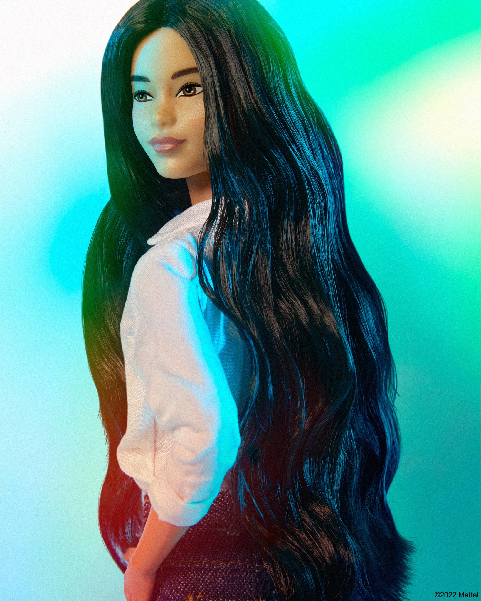 Fashion doll with long black wavy hair, wearing a white shirt and a dark skirt against a vibrant gradient blue and green backdrop.