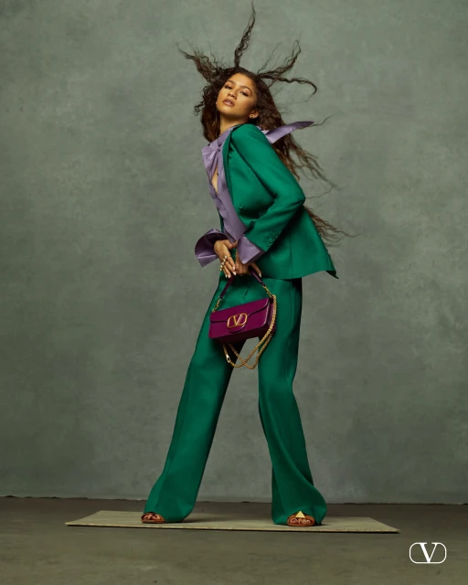 Zendaya in a green suit and purple top holding a purple Valentino bag against a grey backdrop in a fashion editorial shot.