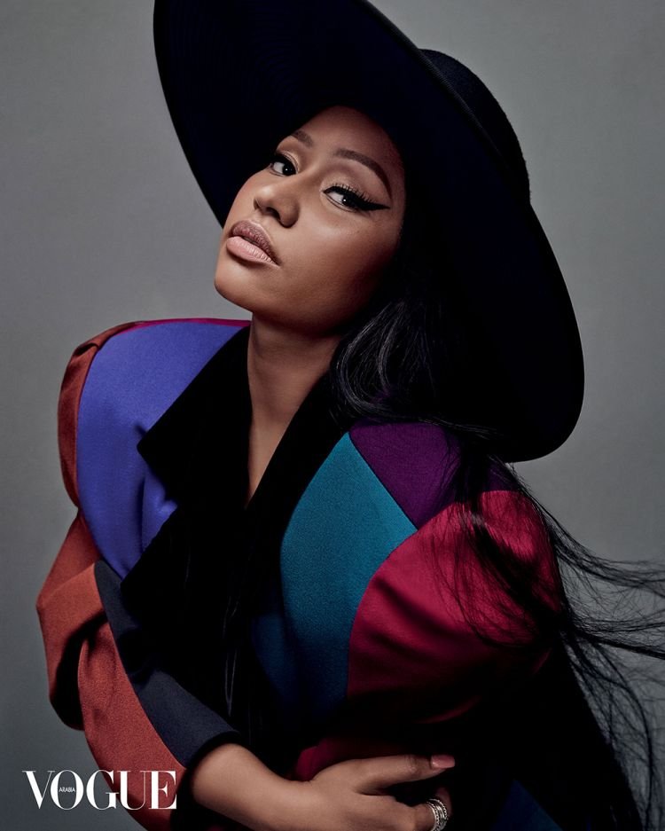 Portrait of Nicki Minaj in a black hat and colorful jacket against a grey backdrop, exuding fashion and celebrity style.