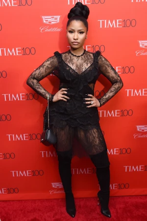 Nicki Minaj poses in a black lace dress and thigh-high boots on a red carpet in front of a TIME 100-branded backdrop.