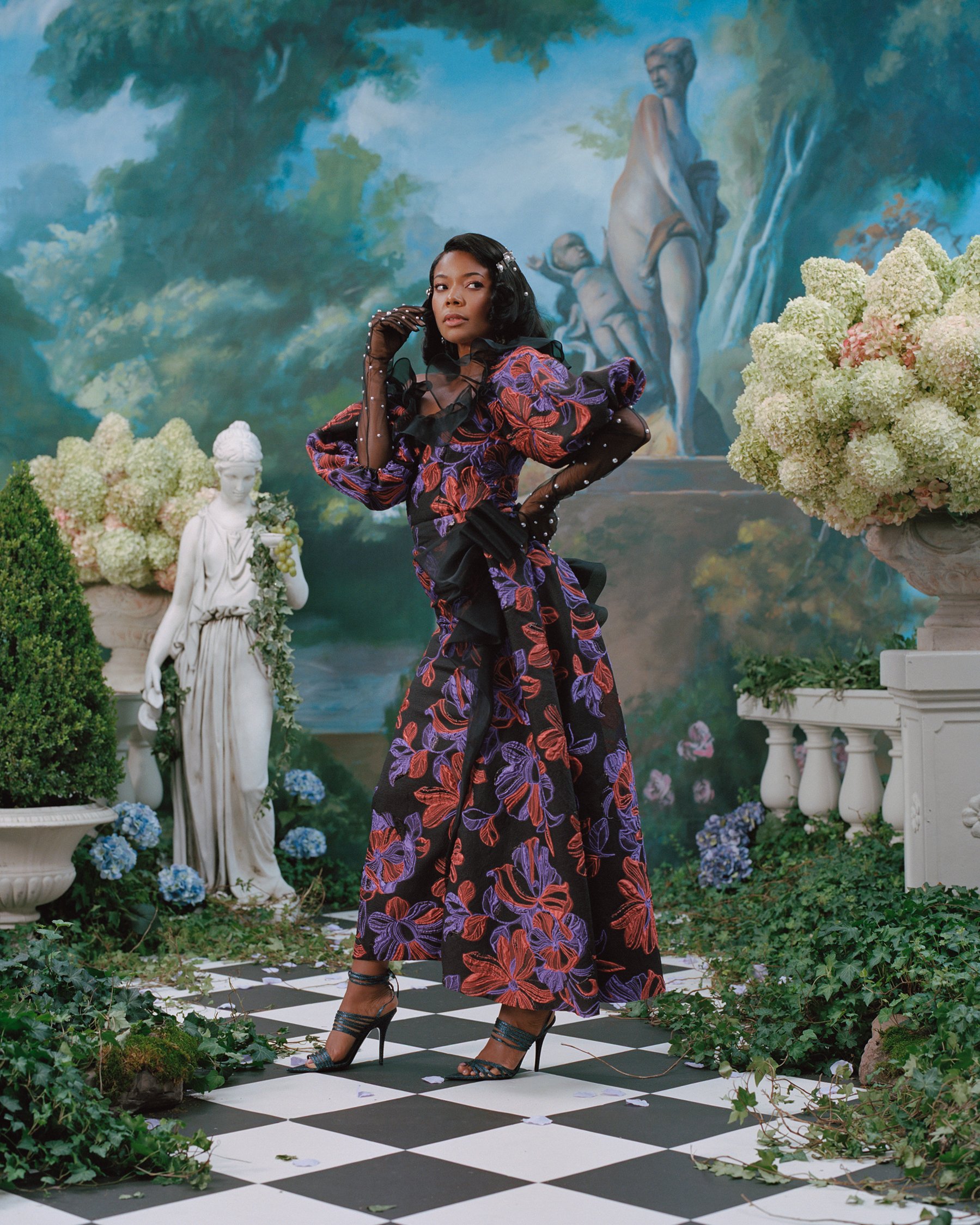 Woman in a floral gown posing in a garden setting with statues and a painted backdrop, wearing gloves and heels on a checkered floor.