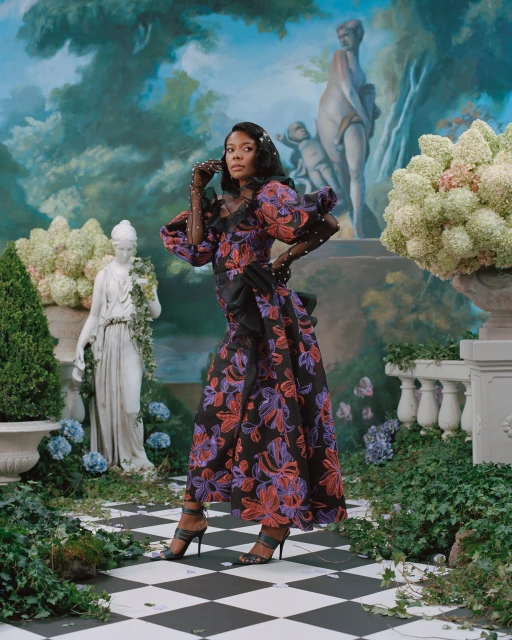 Woman in a floral gown posing in a garden setting with statues and a painted backdrop, wearing gloves and heels on a checkered floor.