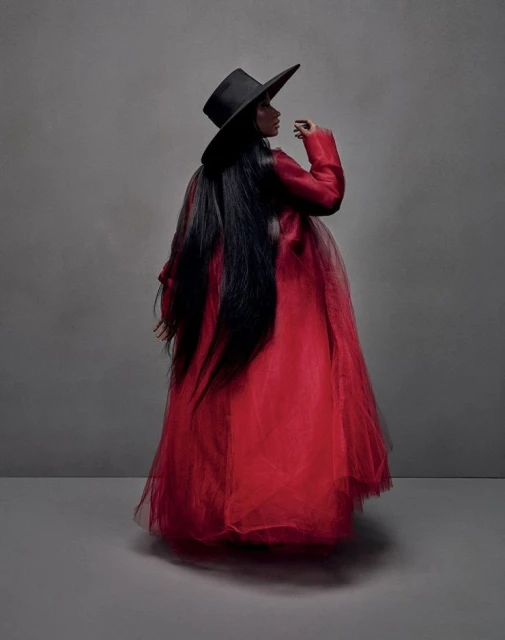 Woman in a red dress and black hat stands in a photo studio with neutral lighting. Long dark hair complements the overall look.