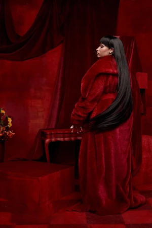 Portrait of an adult female in a red coat and long black hair, posed in a monochromatic red interior setting with a table and flowers.