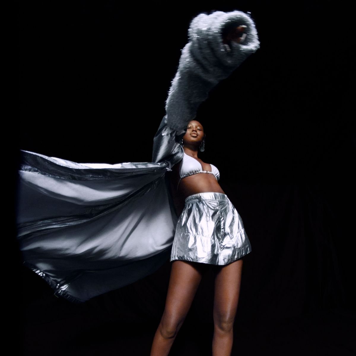 Woman poses in metallic outfit with fur stole against black background. Full shot captures modern fashion and dramatic studio lighting.