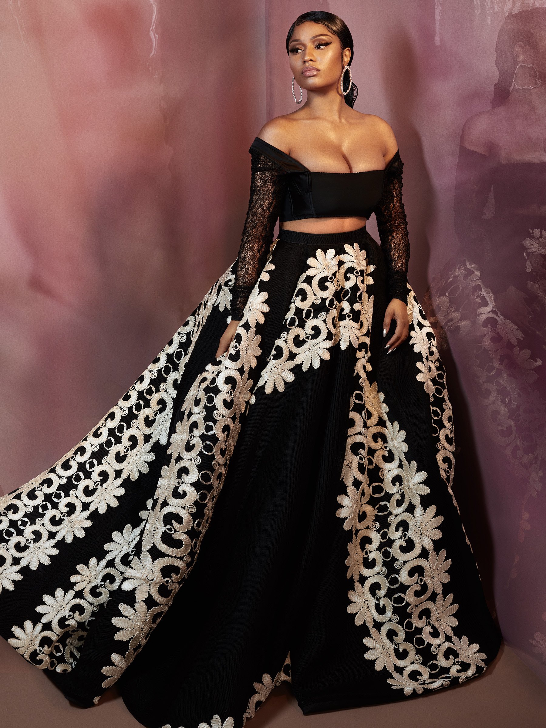 A woman poses in a black two-piece dress with white floral embroidery, hoop earrings, and a plum-colored studio backdrop.