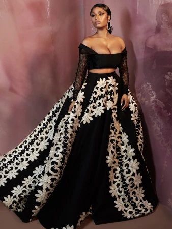 A woman poses in a black two-piece dress with white floral embroidery, hoop earrings, and a plum-colored studio backdrop.