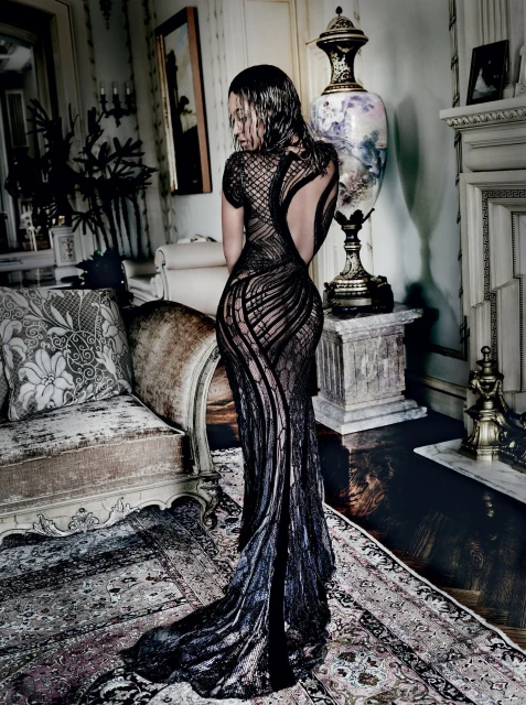 Elegant adult female poses in a backless black lace gown, creating a captivating fashion statement in an ornate interior setting.