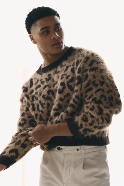 Portrait of a young adult male model wearing a brown and black cheetah-print sweater and beige trousers posing in a studio.
