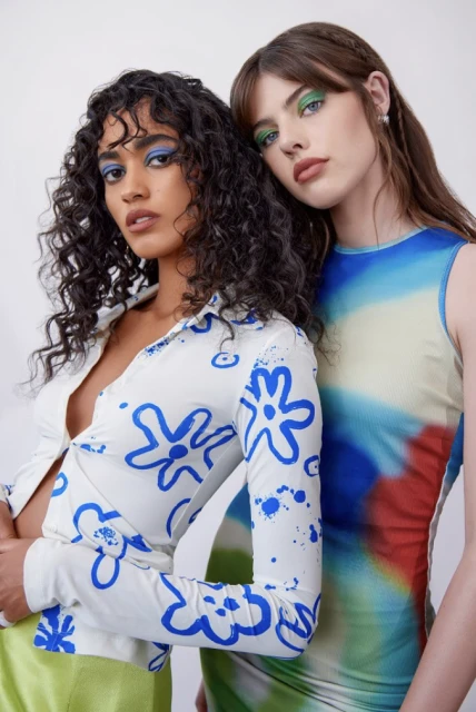 Two fashion models pose in a white photo studio. The model on the left wears a floral top and the model on the right wears a tie-dye dress.