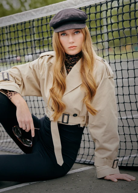 A woman with long wavy red hair poses in a beige trench coat and black leggings in front of a tennis net, holding sunglasses.