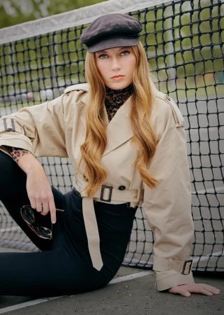 A woman with long wavy red hair poses in a beige trench coat and black leggings in front of a tennis net, holding sunglasses.