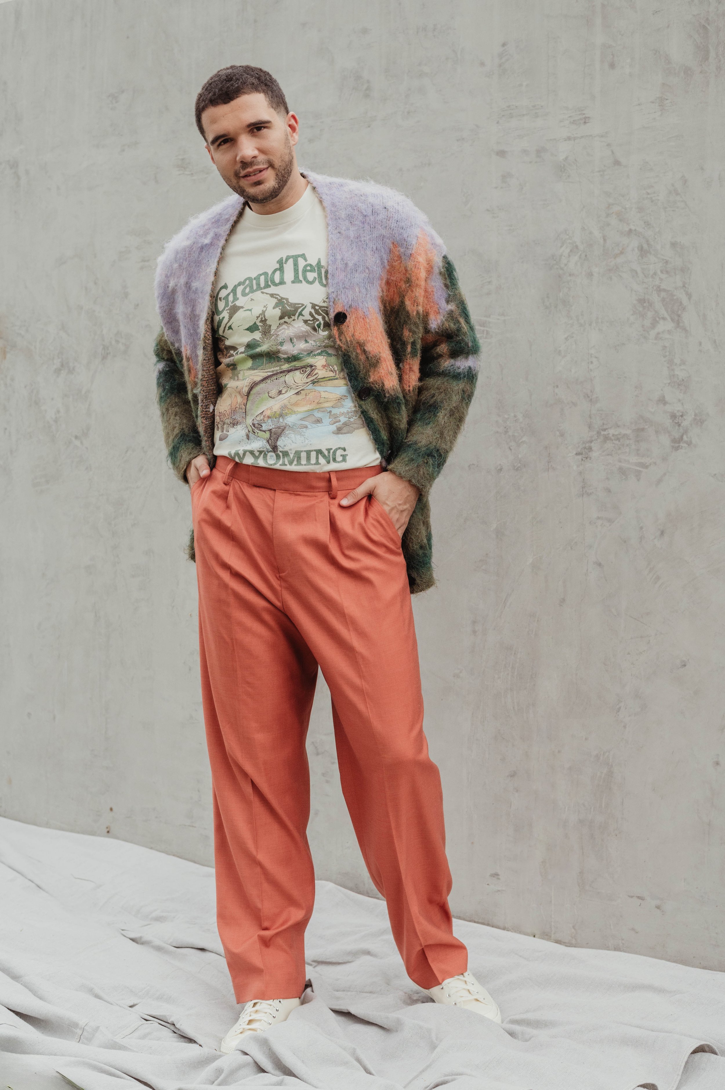 A casually-dressed man stands in front of a textured concrete wall, wearing an illustrated t-shirt, orange pants, and a colorful cardigan.