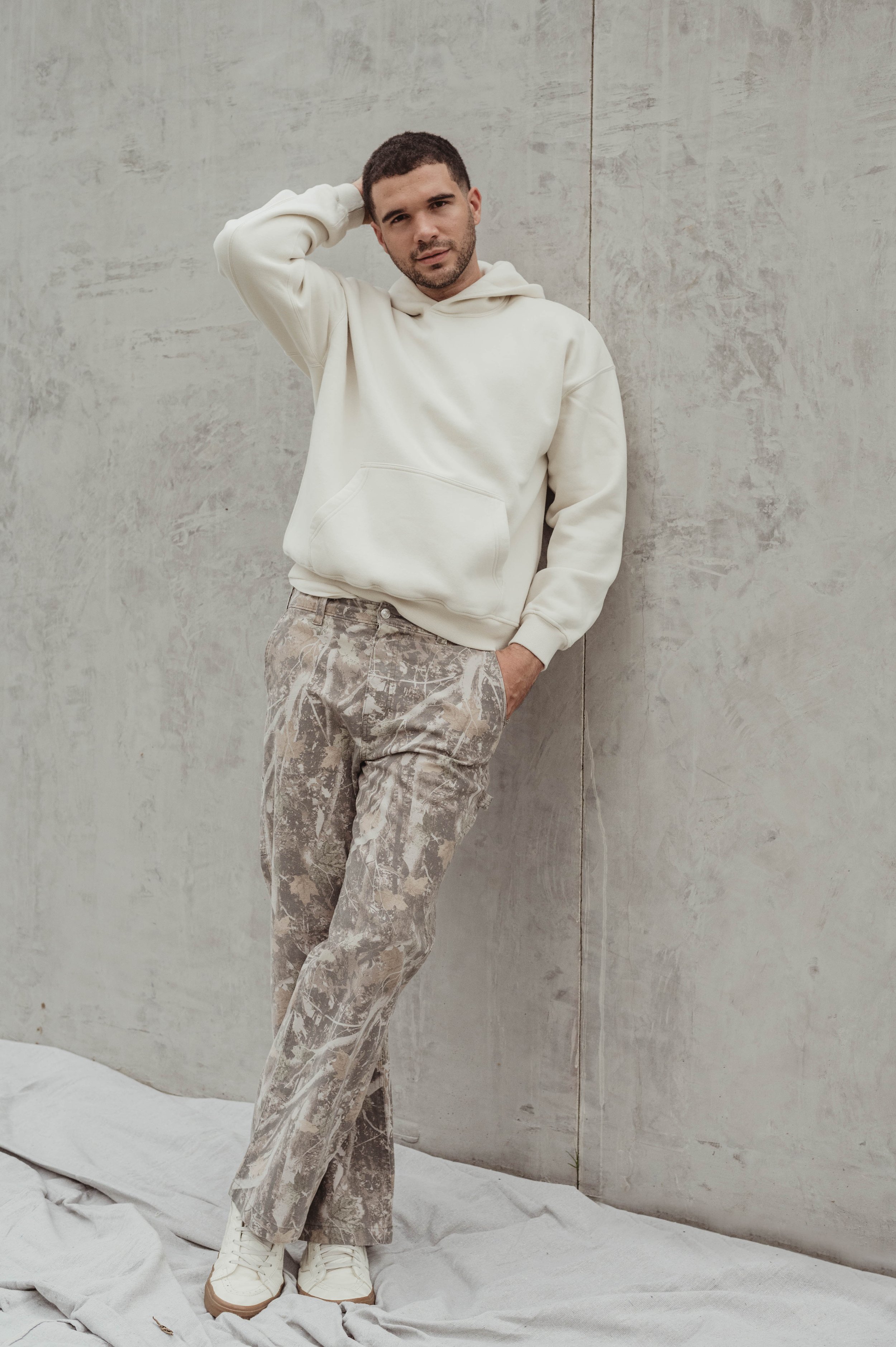 A young man stands against a concrete wall wearing a beige hoodie, camouflage pants, and sneakers in a minimalist fashion portrait.