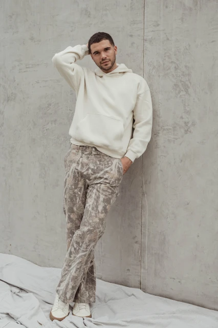 A young man stands against a concrete wall wearing a beige hoodie, camouflage pants, and sneakers in a minimalist fashion portrait.