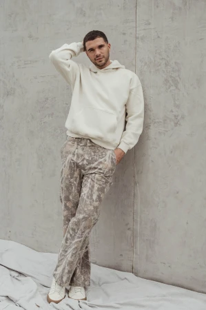 A young man stands against a concrete wall wearing a beige hoodie, camouflage pants, and sneakers in a minimalist fashion portrait.