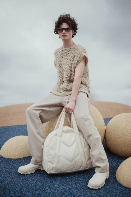 A model wearing a knit sleeveless top and trousers poses with a quilted duffel bag, set against a cloudy sky and patterned terrain.