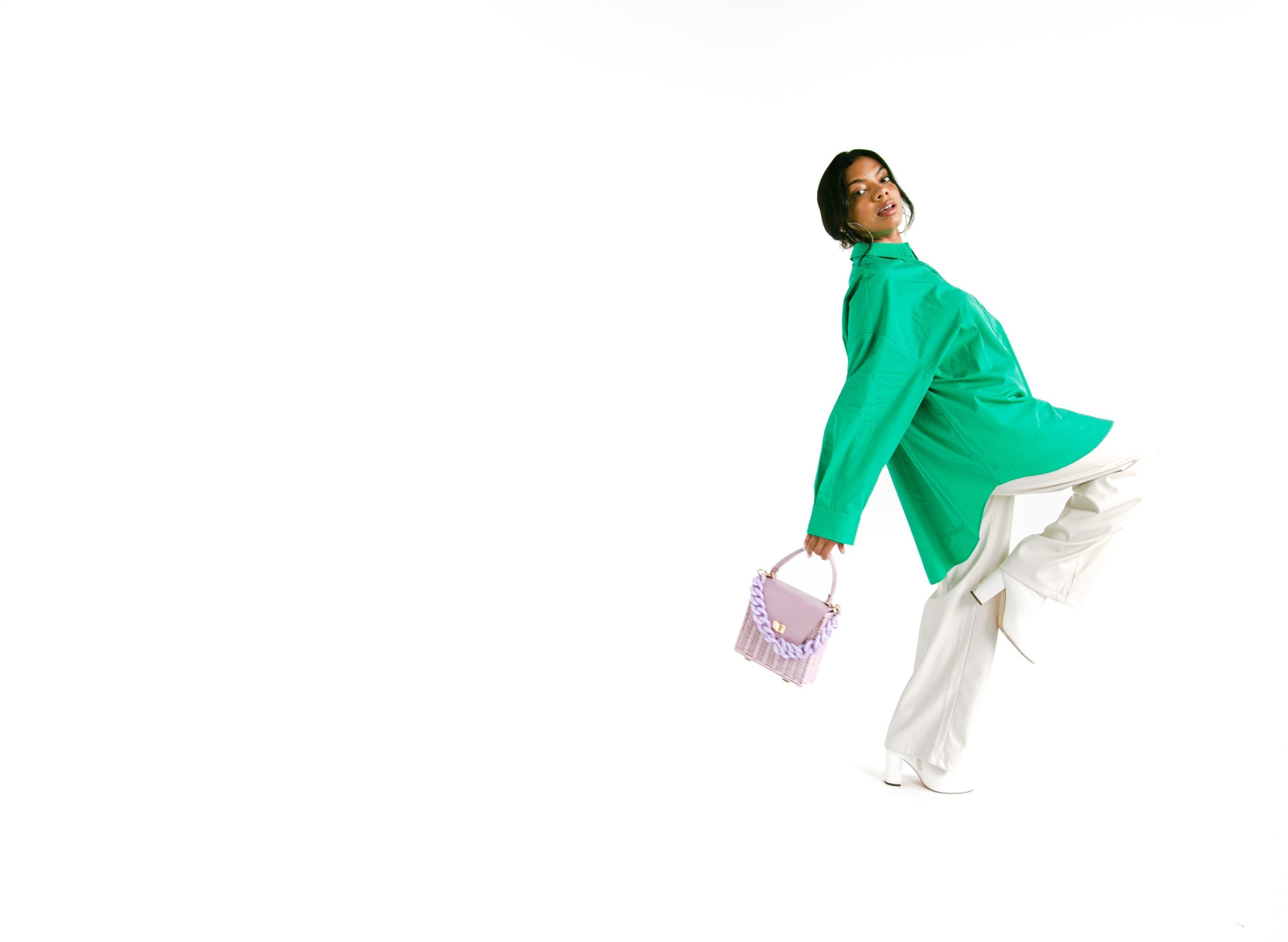 A young woman with short dark hair poses dynamically in a bright green shirt and white pants while holding a small lilac purse.