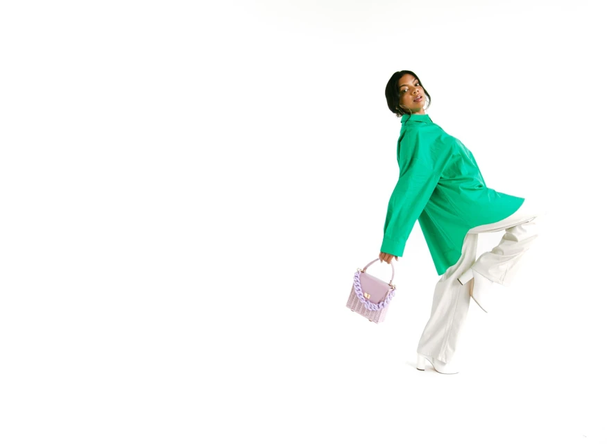 A young woman with short dark hair poses dynamically in a bright green shirt and white pants while holding a small lilac purse.