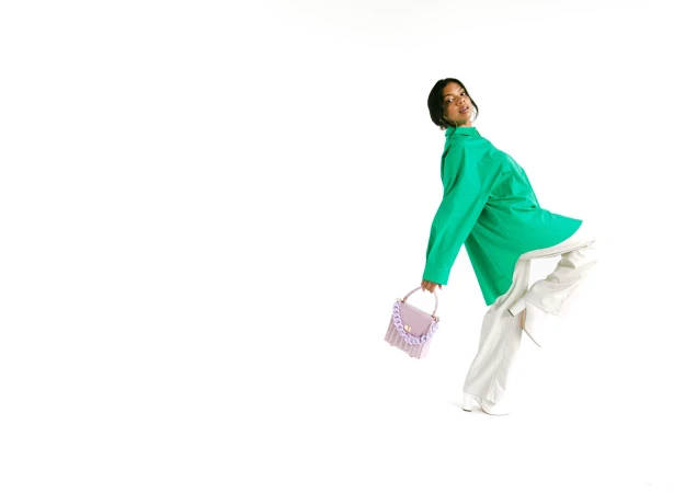 A young woman with short dark hair poses dynamically in a bright green shirt and white pants while holding a small lilac purse.