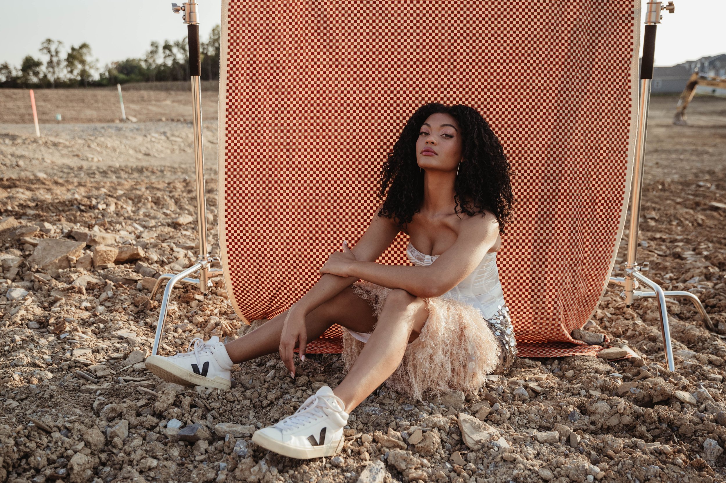 Woman in dress and sneakers poses in an outdoor fashion shoot, with a checkered backdrop and construction site setting at daytime.