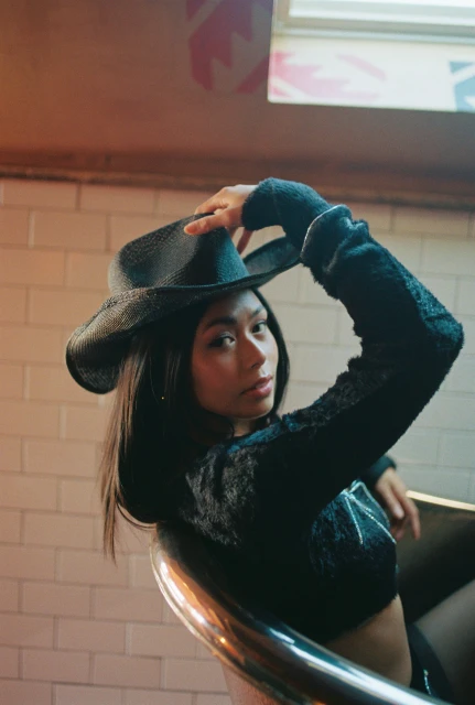 Portrait of woman wearing a black crop top and cowboy hat, seated in front of a white brick wall in an indoor environment.