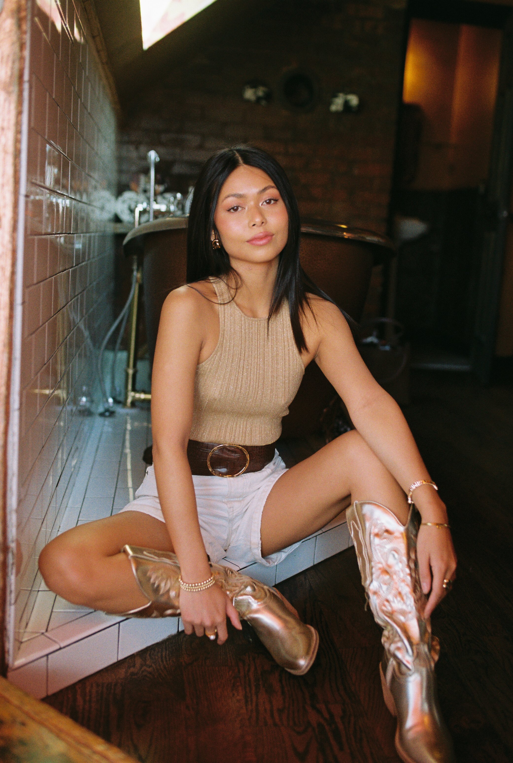 A young woman poses indoors wearing a knit top, shorts, belt, and gold boots, sitting with legs extended toward the viewer, looking at camera.