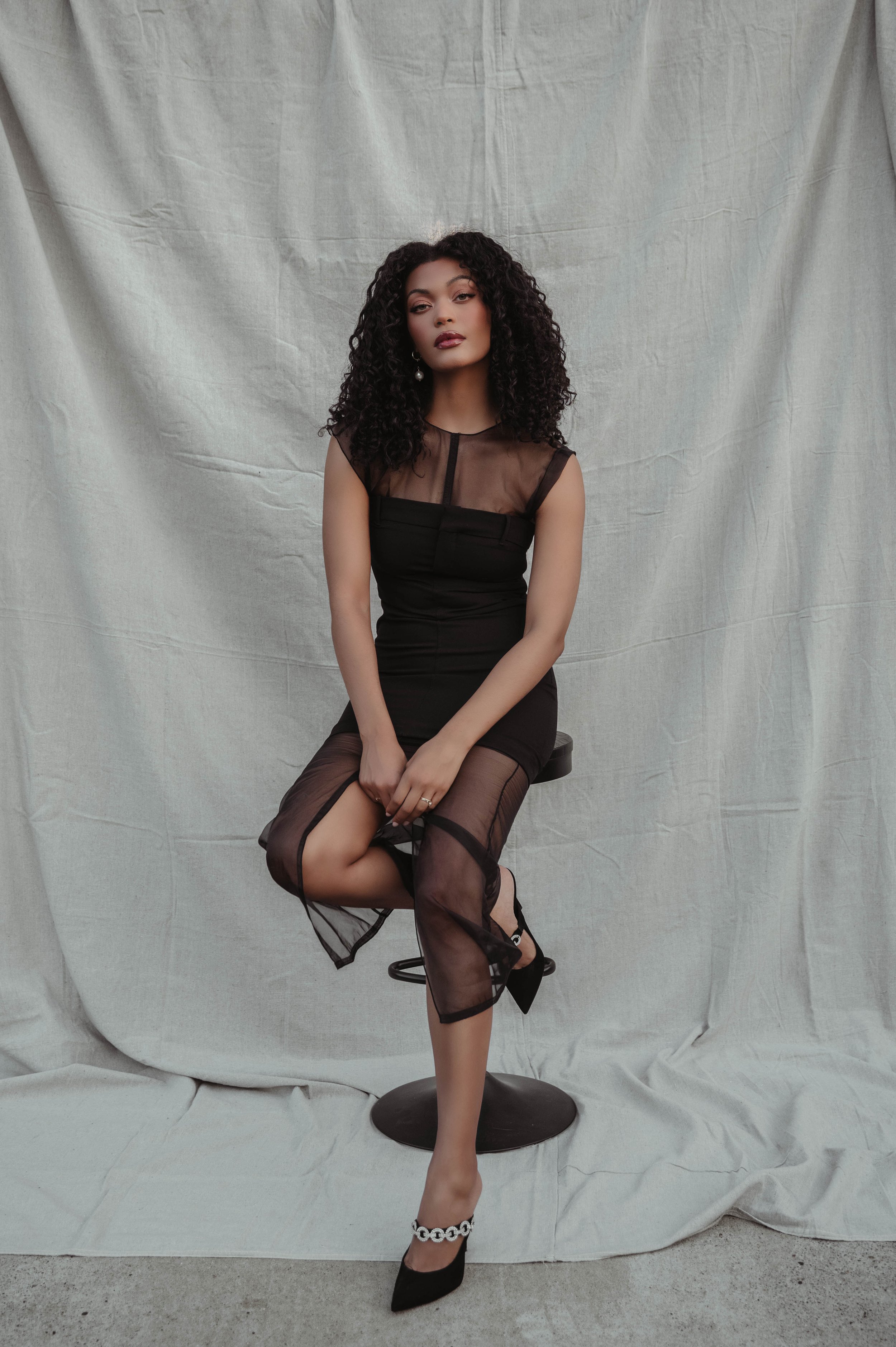 Woman with curly dark hair wearing a black sheer panel dress seated on a stool against a textured white fabric backdrop
